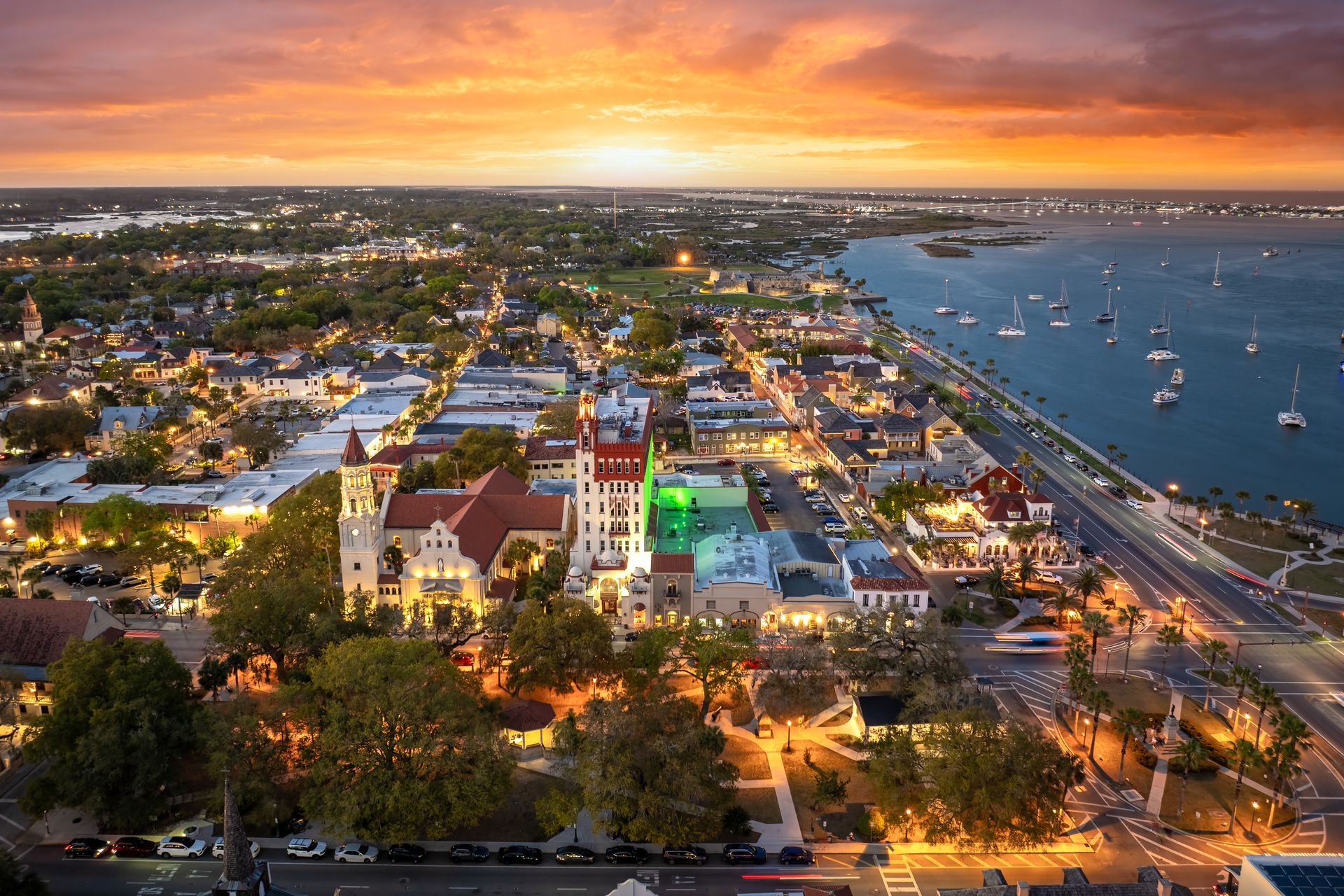 Aerial view of a colorful waterfront town at sunset with boats, roads, and bright city lights