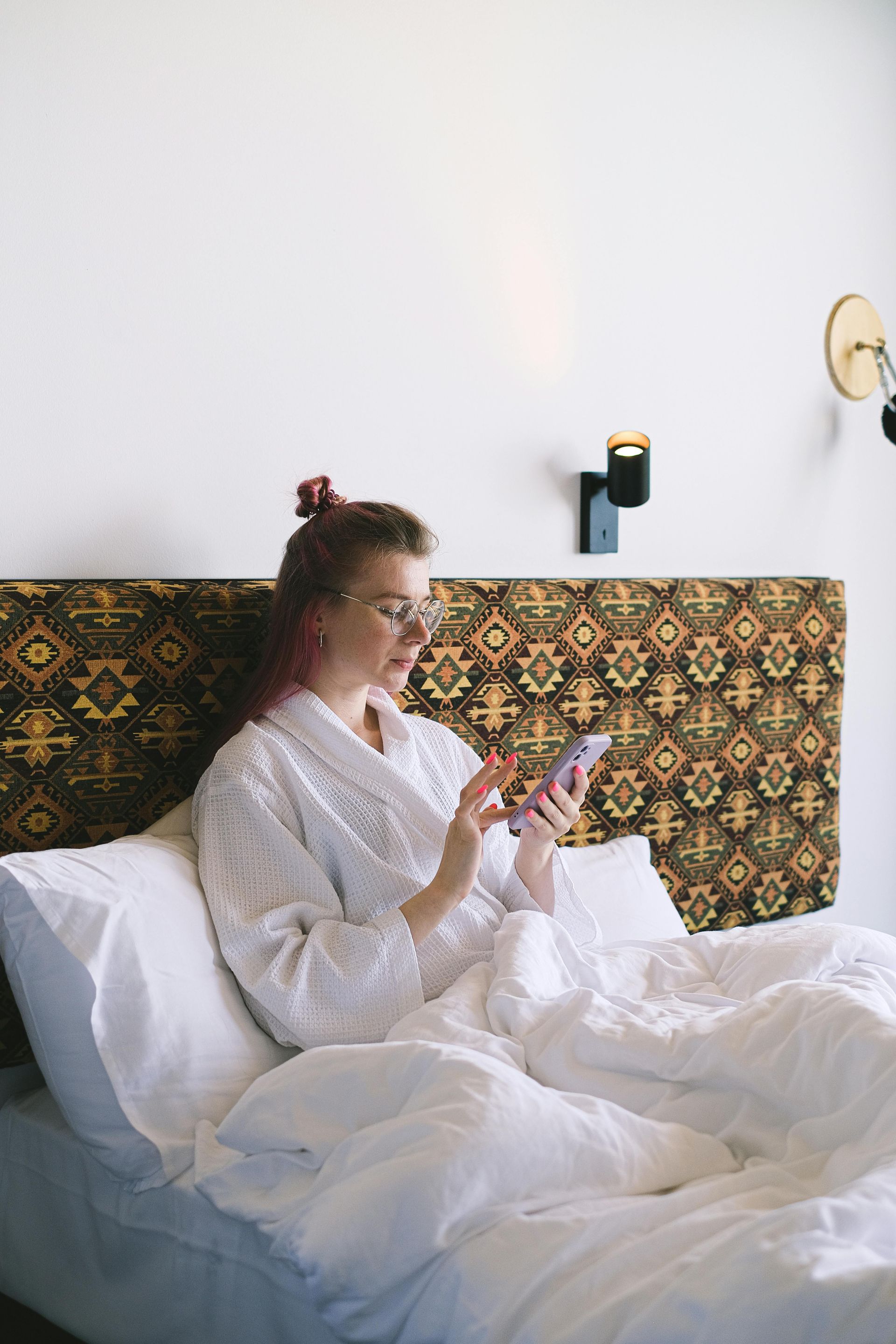 Person sitting in bed in a white robe, looking at a phone in a bright bedroom with patterned headboard