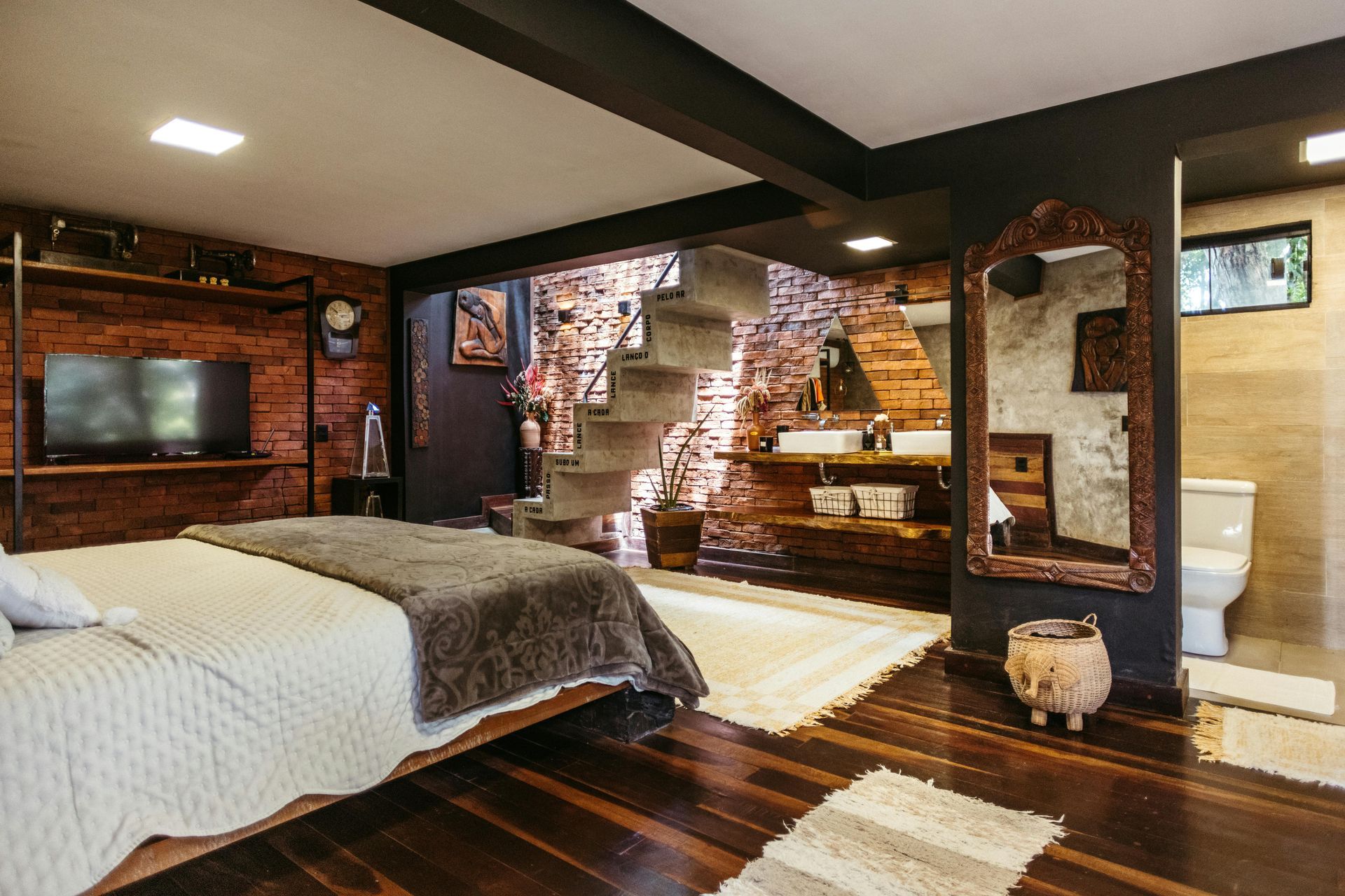 Cozy rustic bedroom with exposed brick, a staircase, bed, TV, and adjacent bathroom.