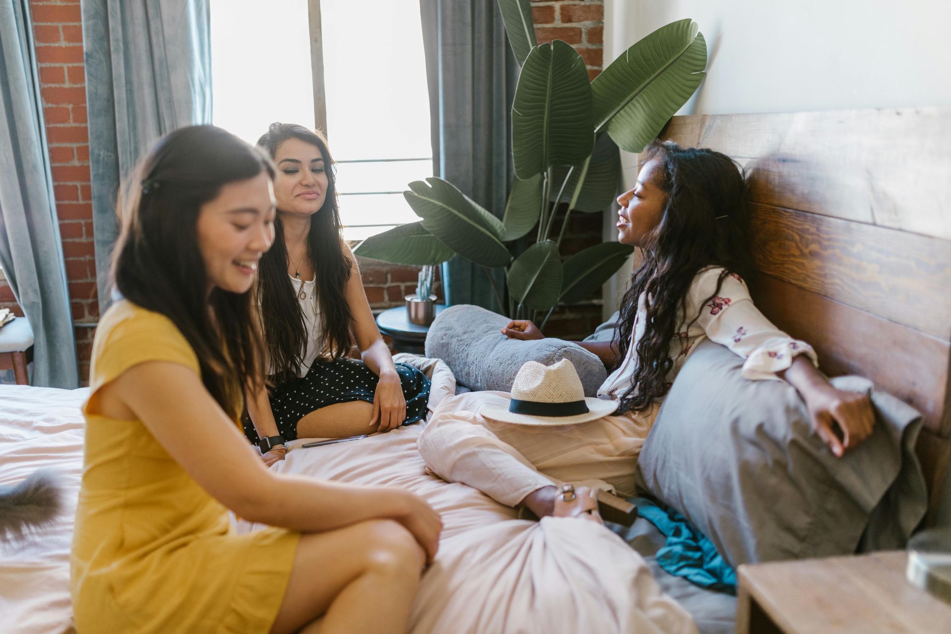 Three friends chatting on a bed in a bright bedroom, with pillows, a hat, and a large plant nearby