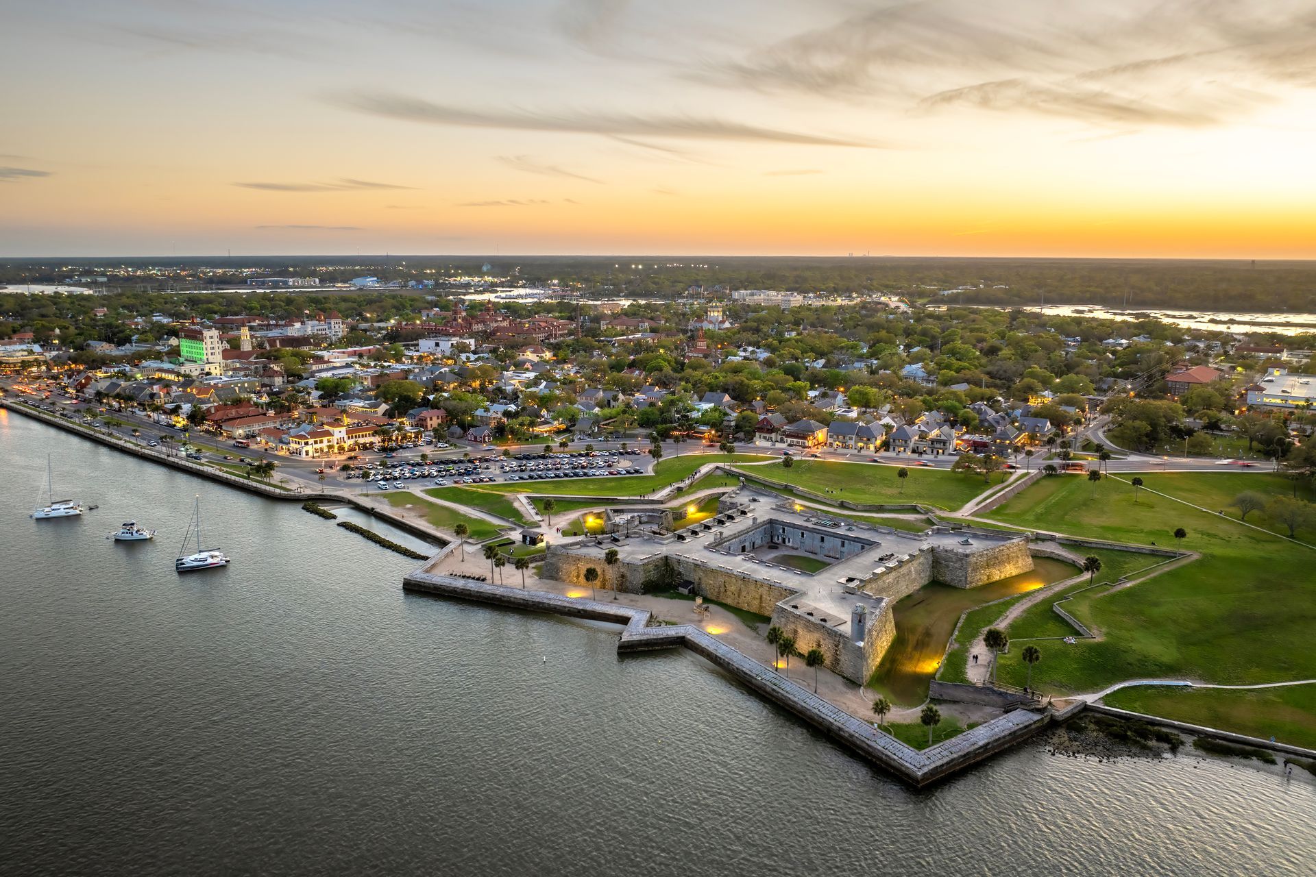 Aerial view of a coastal fort and marina at sunset, with boats and a town along the shoreline