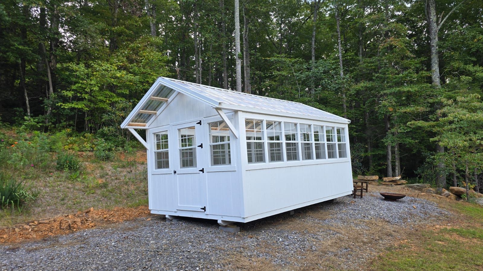 White greenhouse with windows stands on gravel, surrounded by trees.