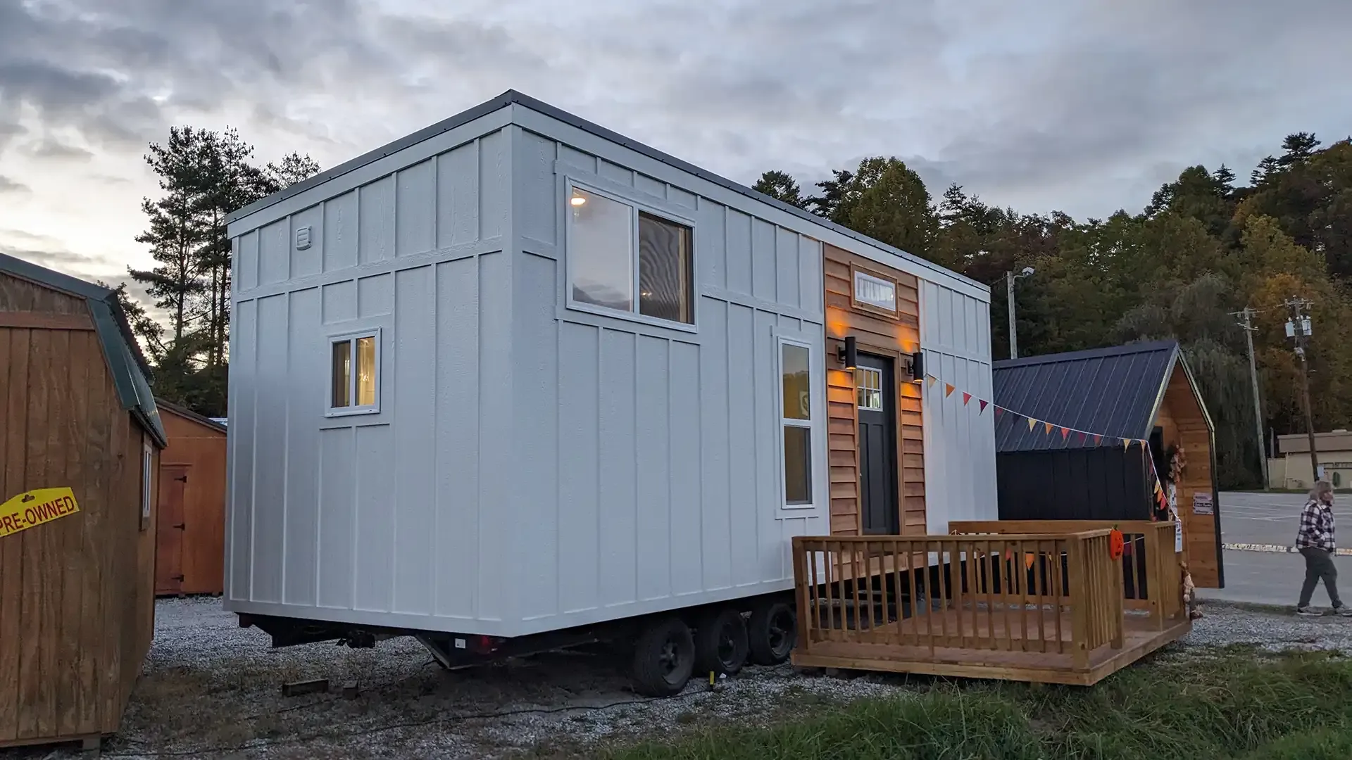 White tiny house with wooden deck, parked on gravel, cloudy day.