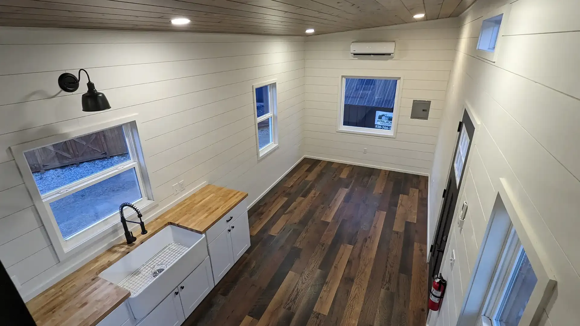 Interior of a tiny home with a kitchen, wood floor, white walls, and windows.