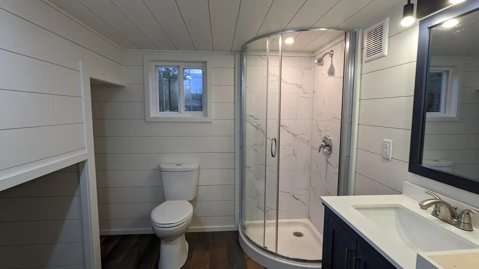 Bathroom with white paneled walls, curved shower, toilet, vanity, and small window.