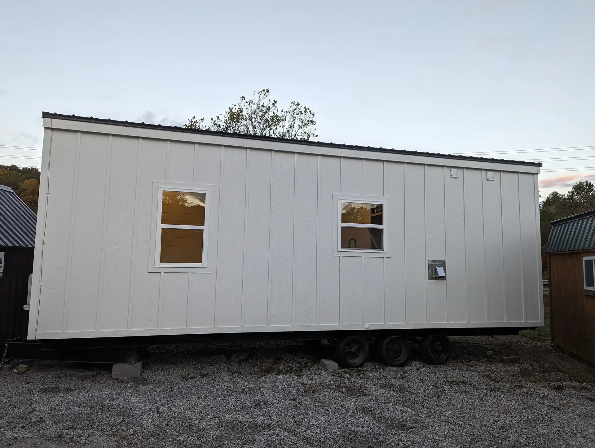 White tiny home on trailer with two windows, parked on gravel lot.
