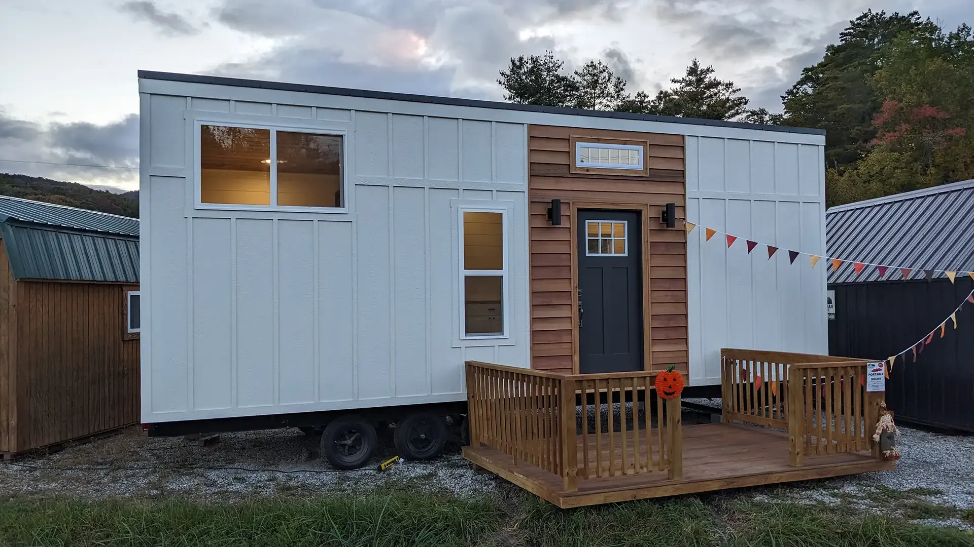 Tiny house on a trailer, white exterior, small porch, wooden accents, gray door, lit window, daytime.