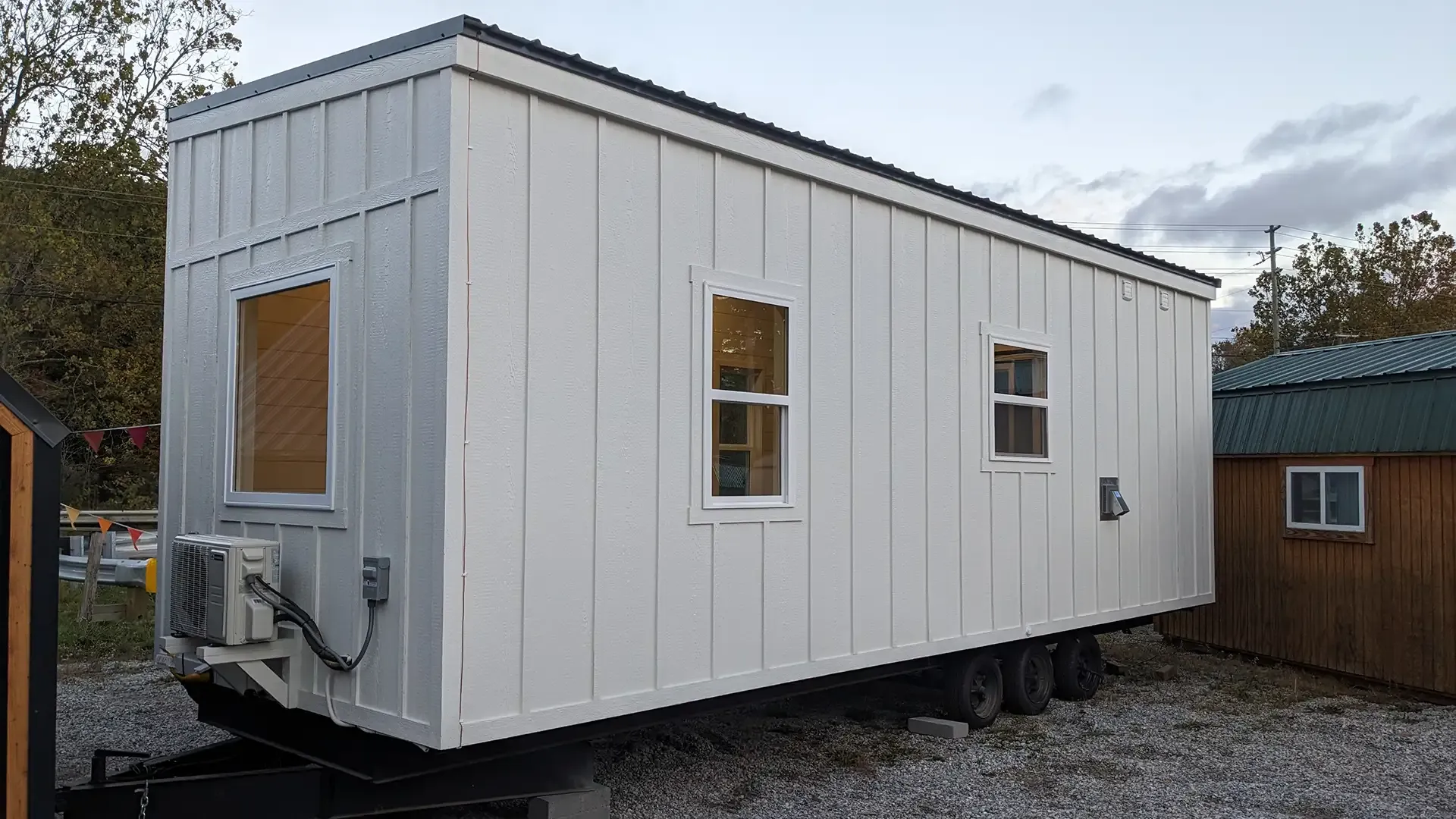 White tiny house on a trailer with a dark roof and three windows, parked outside.
