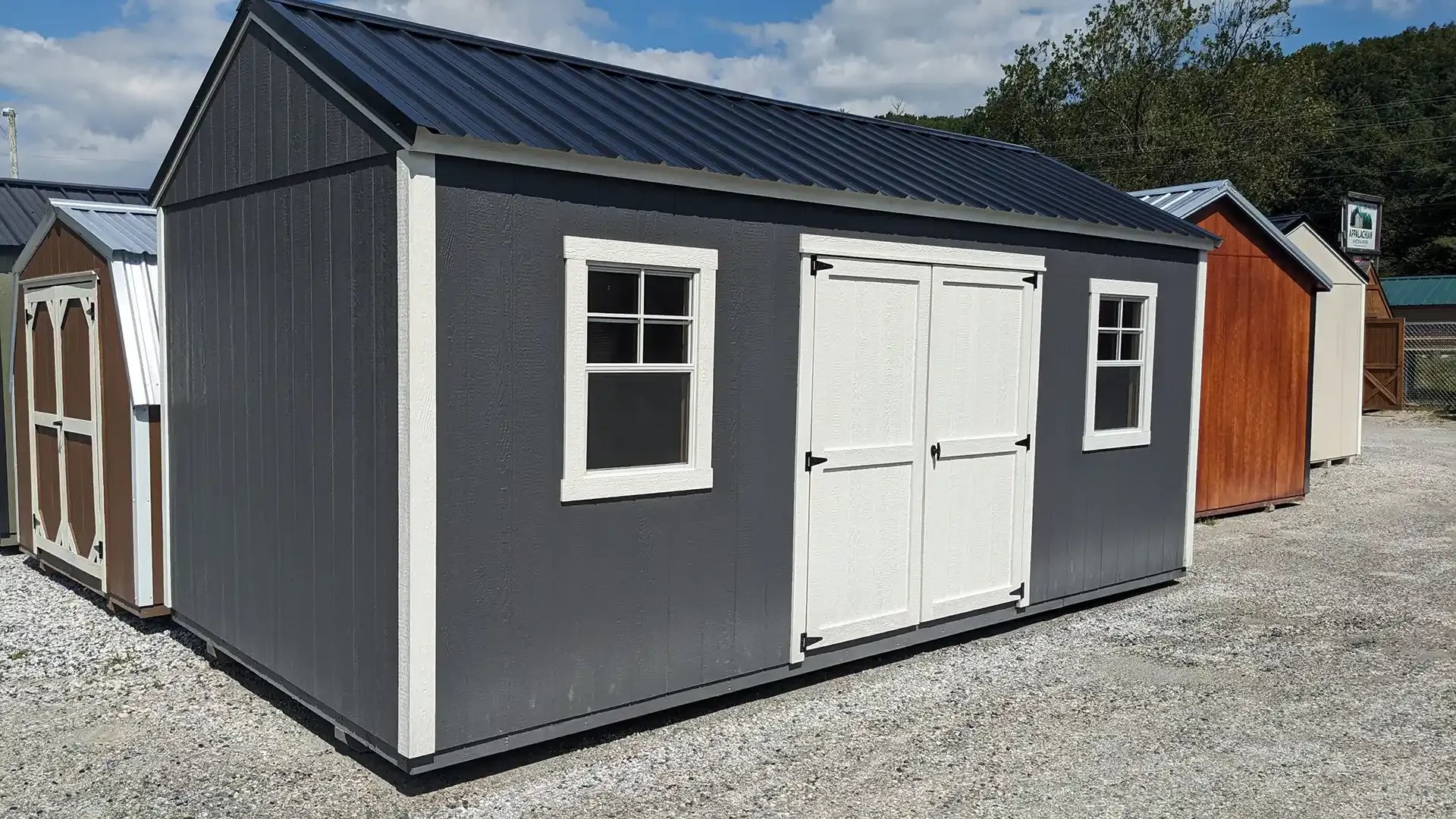 Gray storage shed with white trim, doors, and windows, set outside under a blue sky.