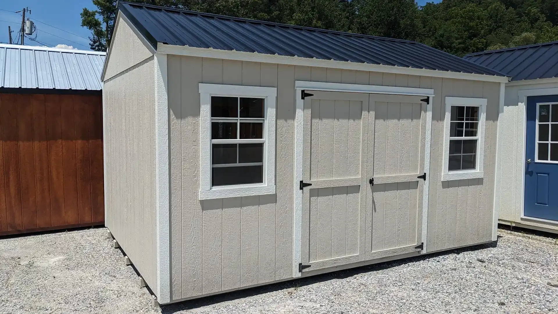 Tan shed with black roof, white trim, and double doors. Two windows flank the doors.