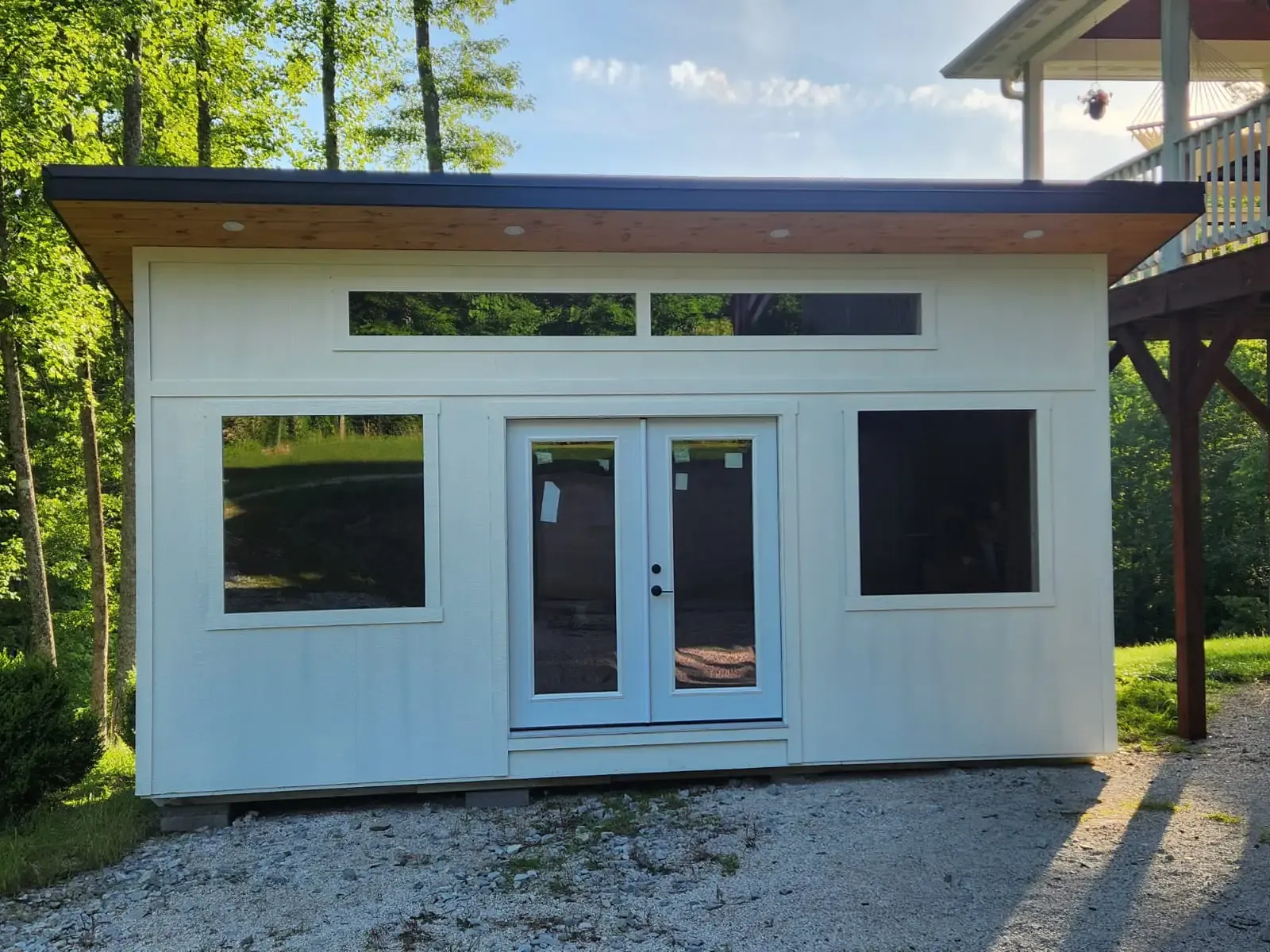 White modern shed with black trim, glass windows, and double doors in a gravel driveway.