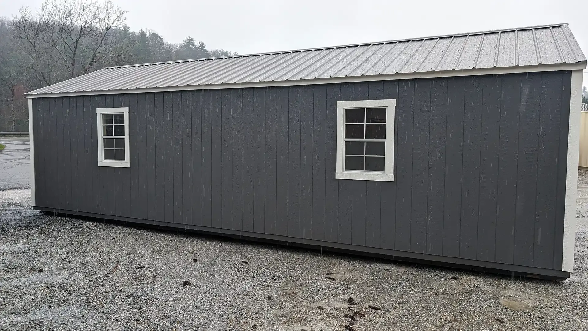Dark gray shed with two white-framed windows, a metal roof, and white trim, sitting on gravel.