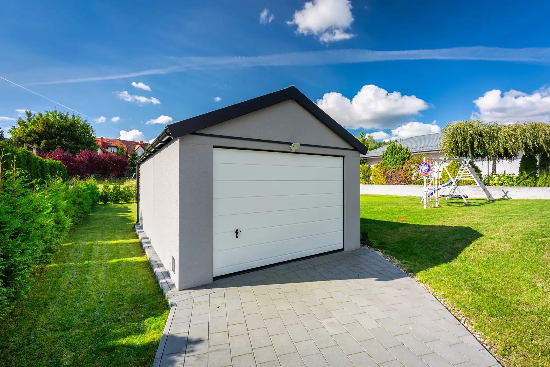 Grey garage with white door on a sunny day, surrounded by green grass and a paved driveway.