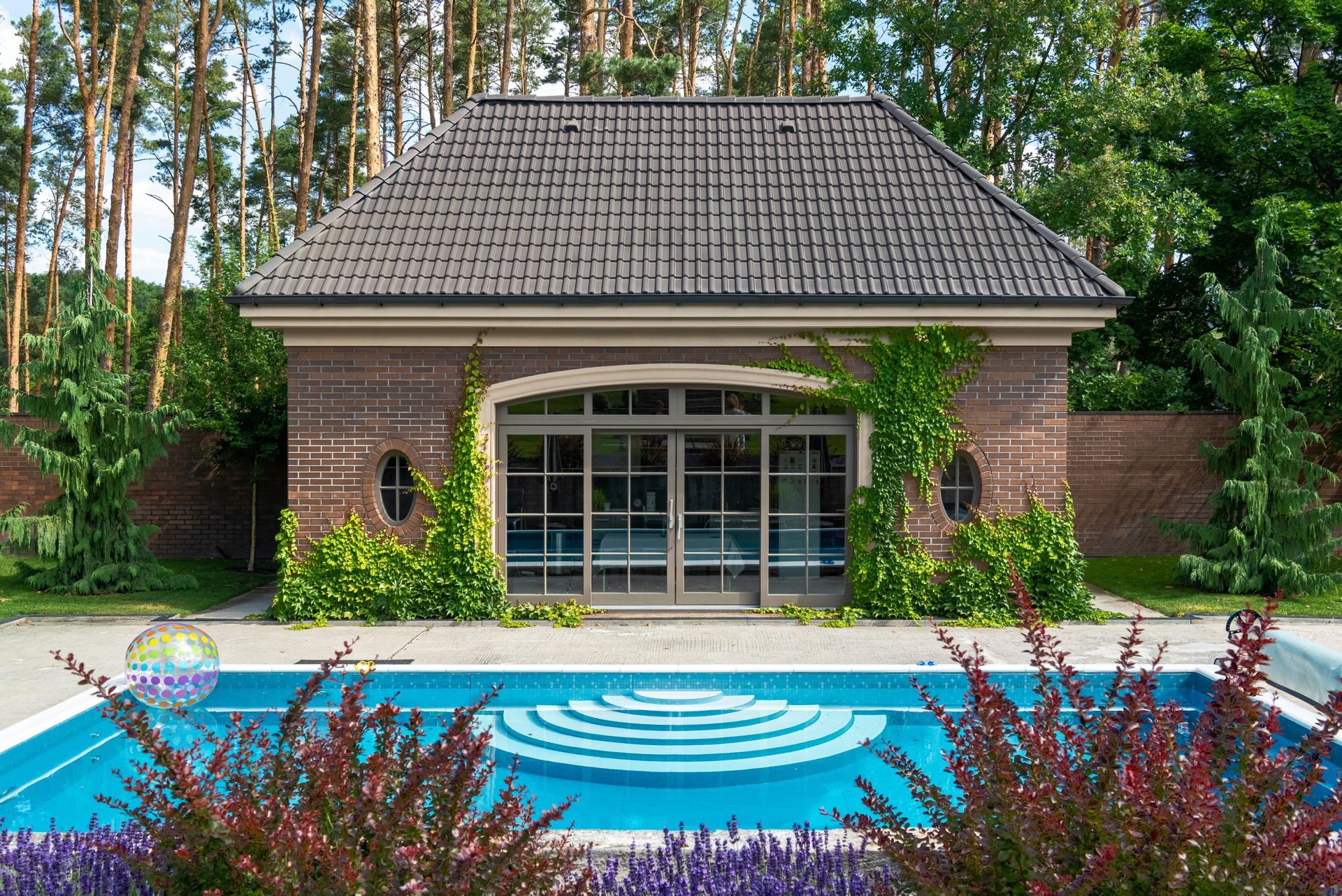 Swimming pool with building in the background, brick, brown roof, green foliage, clear blue water.