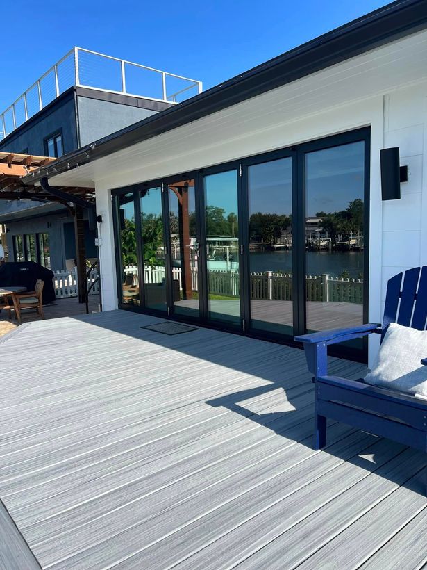 Modern waterfront patio with glass doors, white deck boards, and blue chairs under clear sunlight