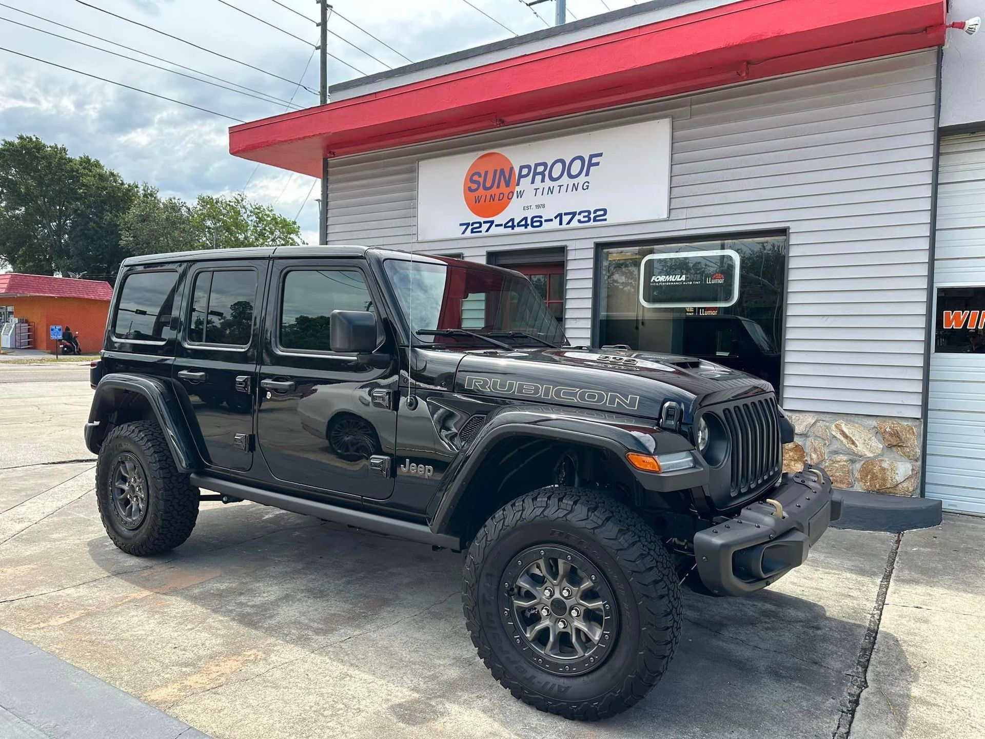 Black Jeep Wrangler parked outside an auto shop with a red awning and sign.