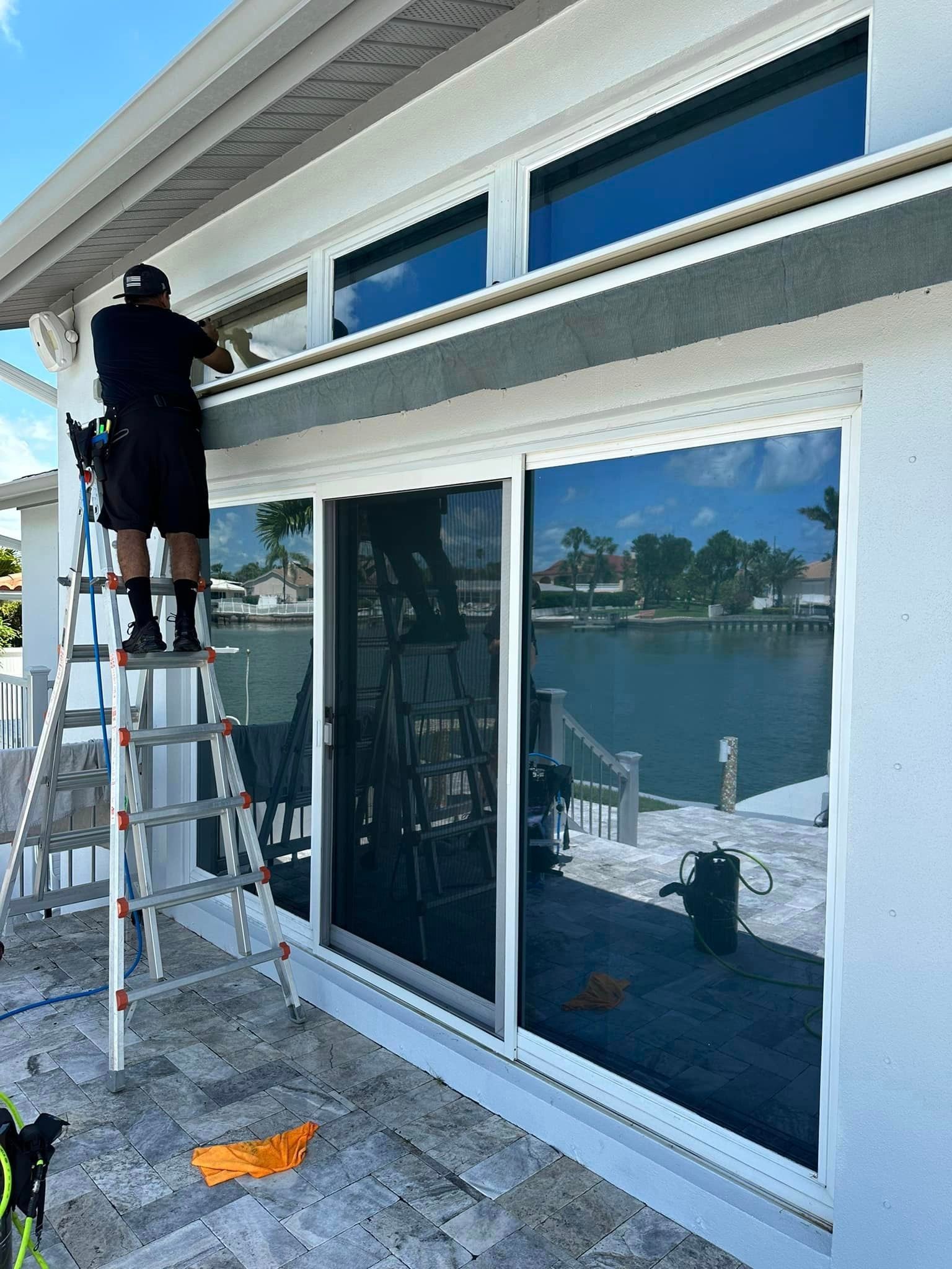 Worker on ladder painting exterior trim above sliding glass doors on a white house.
