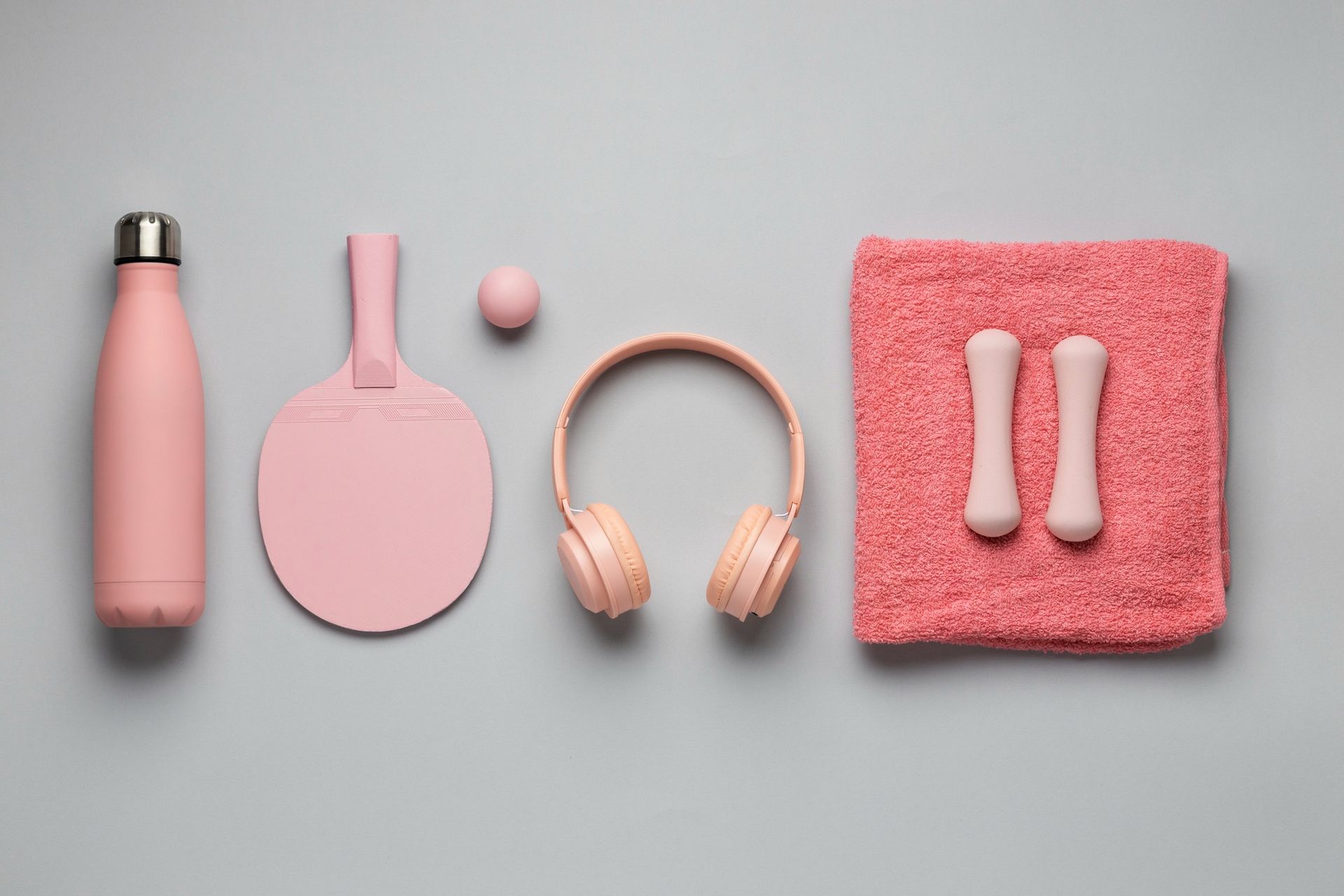 A flat-lay of light pink fitness gear, including a water bottle, ping-pong paddle, ball, headphones, towel, and weights.