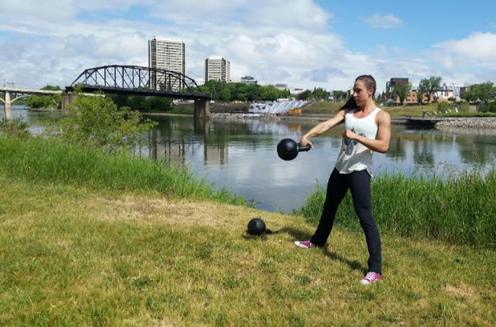 A person exercises with kettlebells on a grassy riverbank near a bridge and city buildings.