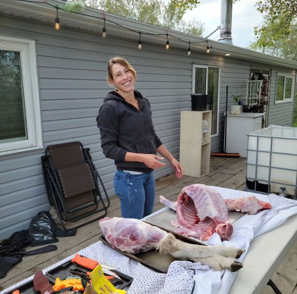 A person stands smiling behind a table outdoors on a deck, preparing raw butchered game meat on trays.