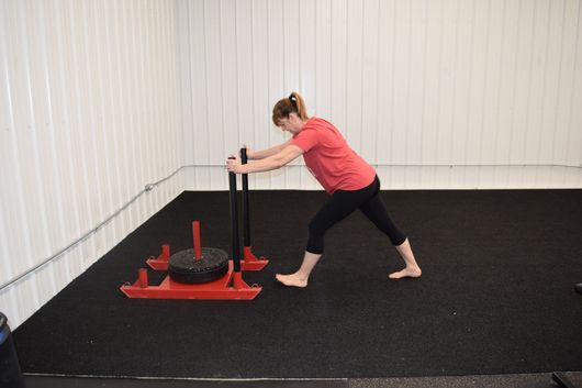 A person wearing a red shirt and black leggings barefoot pushes a red weight sled across a black gym floor.