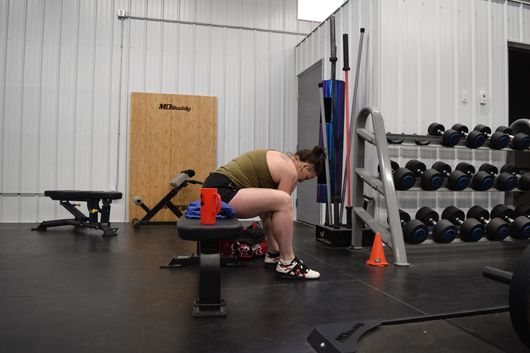 A person in athletic gear sits on a gym bench with head bowed in a brightly lit fitness studio.