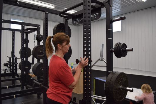 A person in a red shirt adjusts a rack attachment in a gym with weights, while a child sits on the floor nearby.