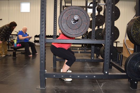A person in a red shirt performs a barbell squat inside a gym power rack while two others watch from a nearby bench.