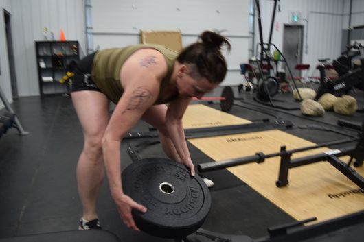 A person in a gym leans over to load a black weight plate onto a barbell positioned on a landmine attachment.