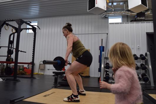 A person wearing a green tank top and black shorts performs a barbell deadlift while a child watches in a gym.