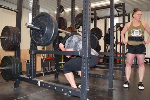 A person performs a squat with a barbell in a rack while another person stands nearby and observes in a gym.