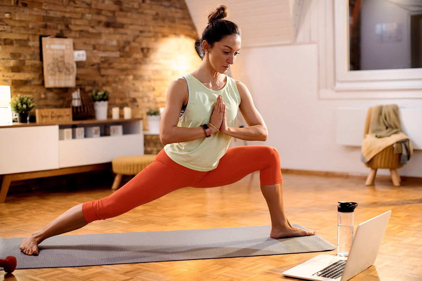 A person in a light tank top and orange leggings practices a yoga side lunge while watching a laptop at home.