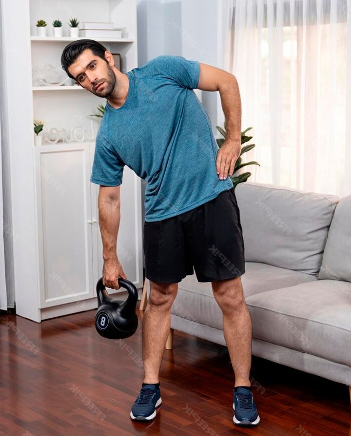 A person in a blue t-shirt stands in a living room, performing a weighted side bend exercise with a black kettlebell.