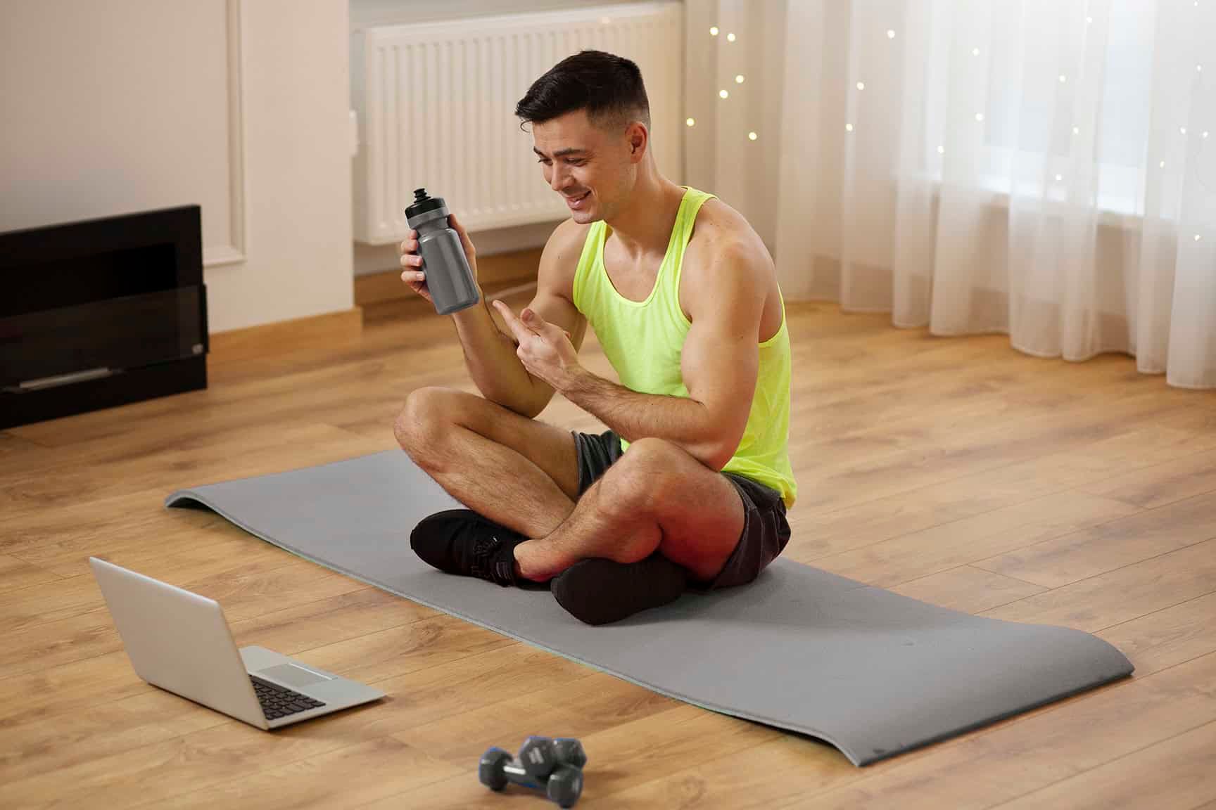 A person in a neon yellow tank top sits on a yoga mat, holding a water bottle while looking at a laptop.