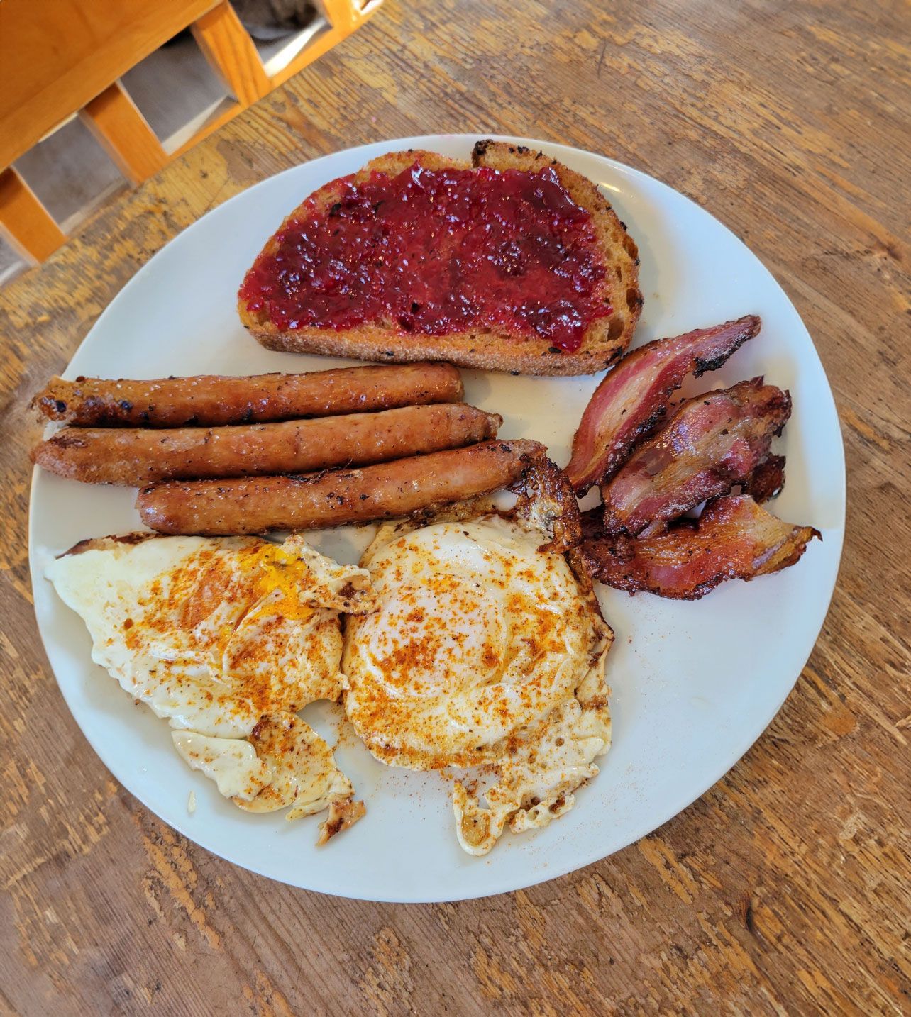 A plate of breakfast featuring two fried eggs, three sausages, several strips of crispy bacon, and toast with jam.
