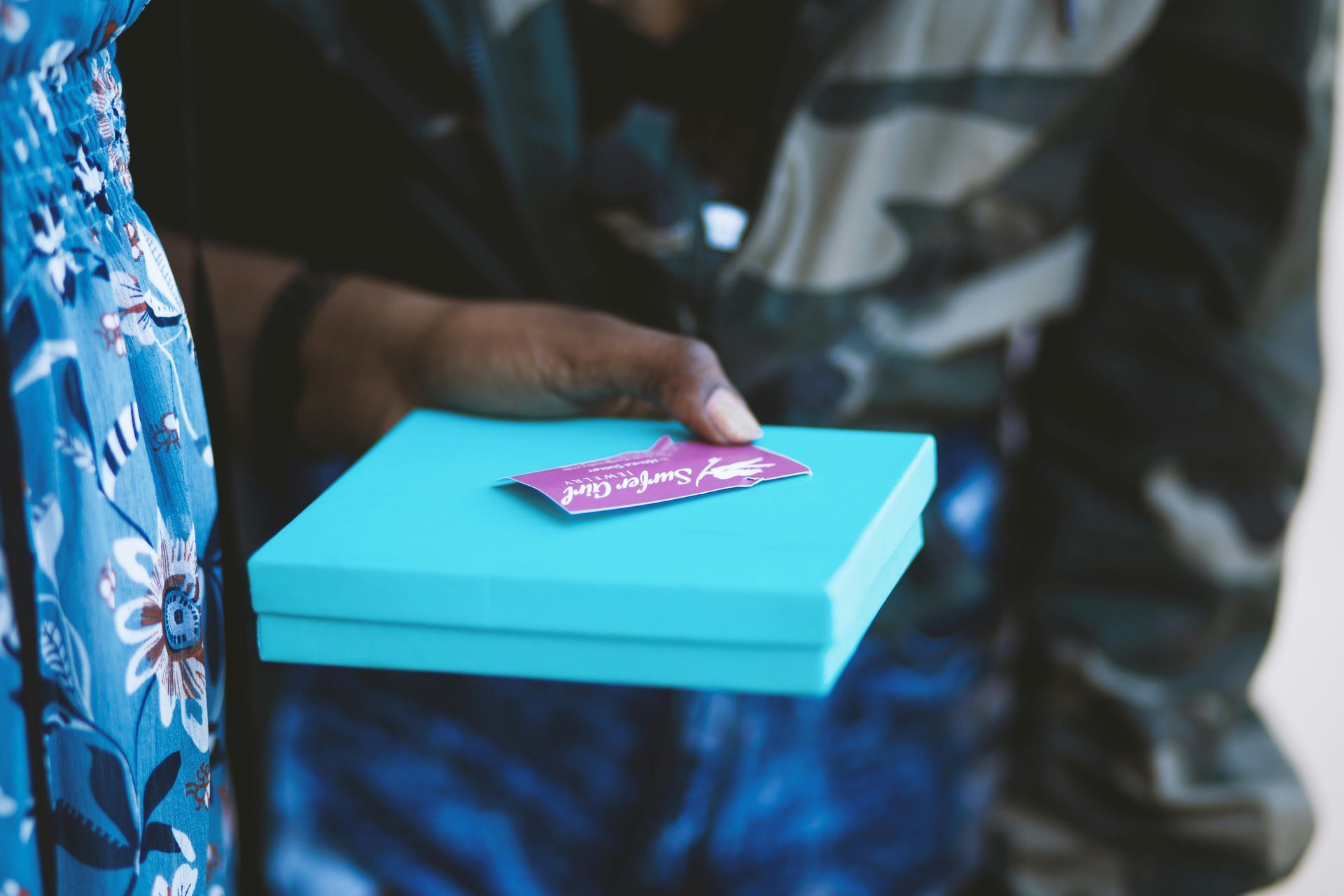 A person’s hand holds a small, light blue gift box topped with a purple tag.