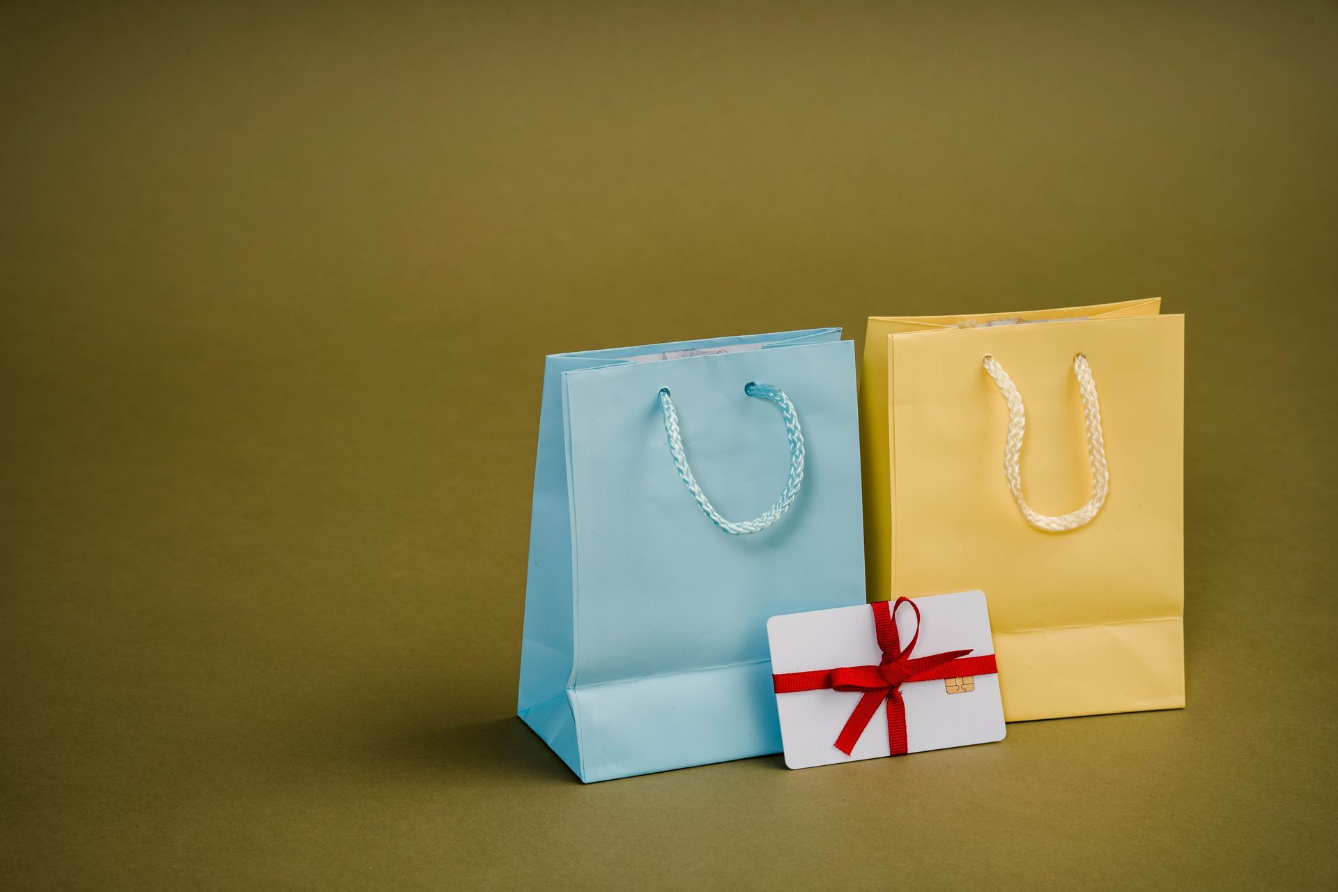 A light blue bag and a yellow bag stand next to a white gift card tied with a red ribbon against a dark green background.