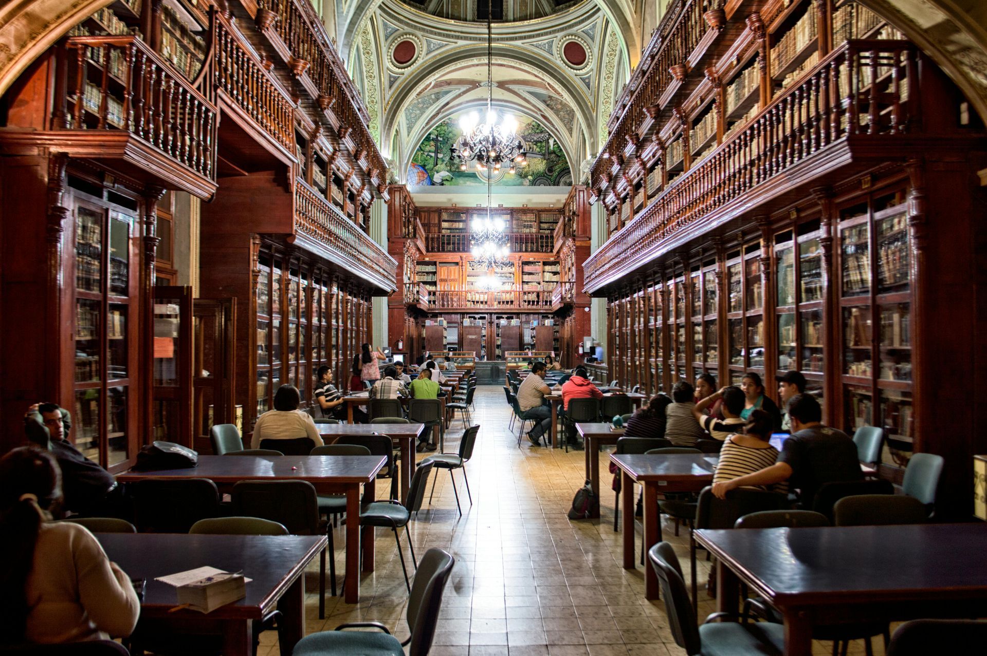 People study at tables in a grand, wood-paneled library featuring high, arched ceilings and multi-level bookshelves.