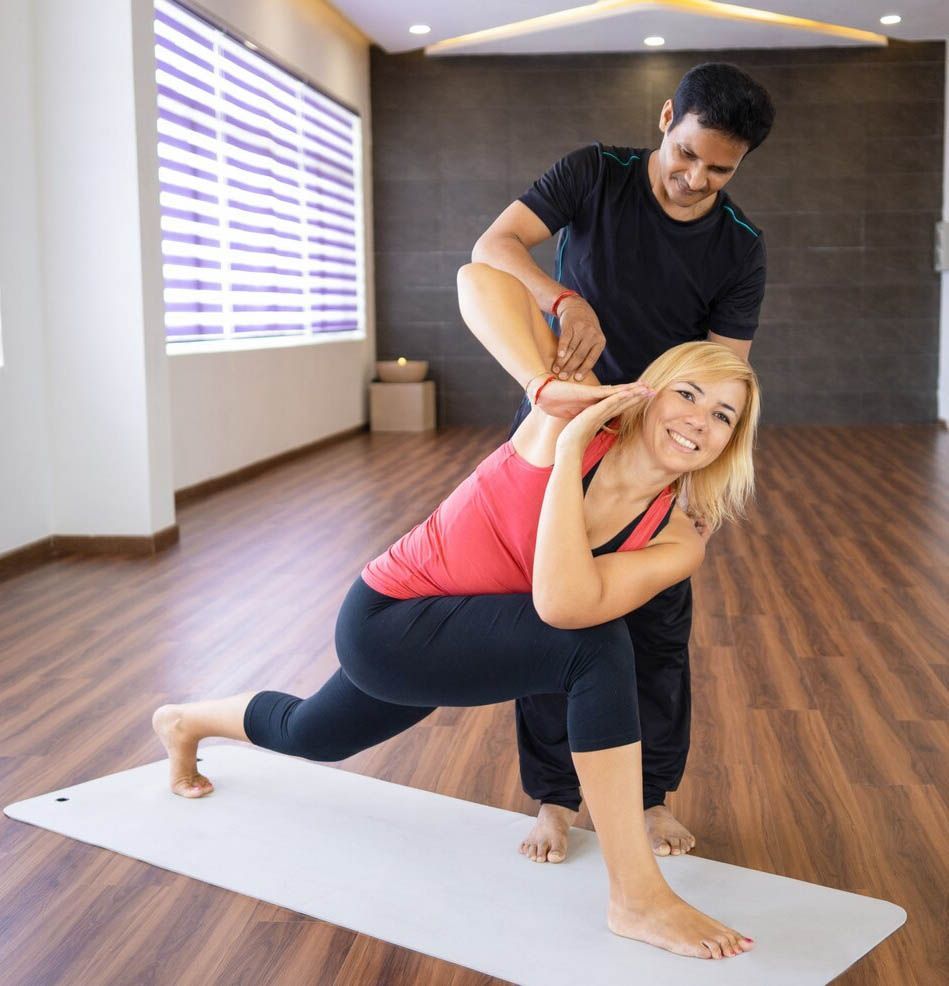 An instructor guides a smiling person in a yoga lunge with a torso twist on a mat in a studio.