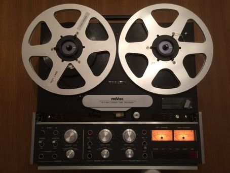 Reel-to-reel tape recorder with two silver reels. Black body, knobs, and lit VU meters.