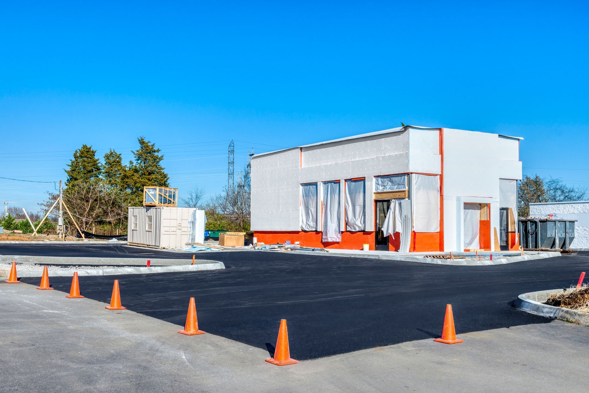 Exterior view of a building under construction, white walls with orange accents, new asphalt, and orange cones.