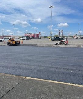 Freshly paved asphalt with construction equipment, and a distant commercial building on a cloudy day.