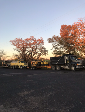 Construction trucks on a road at dusk. A black dump truck and two yellow trucks with trailers are visible.