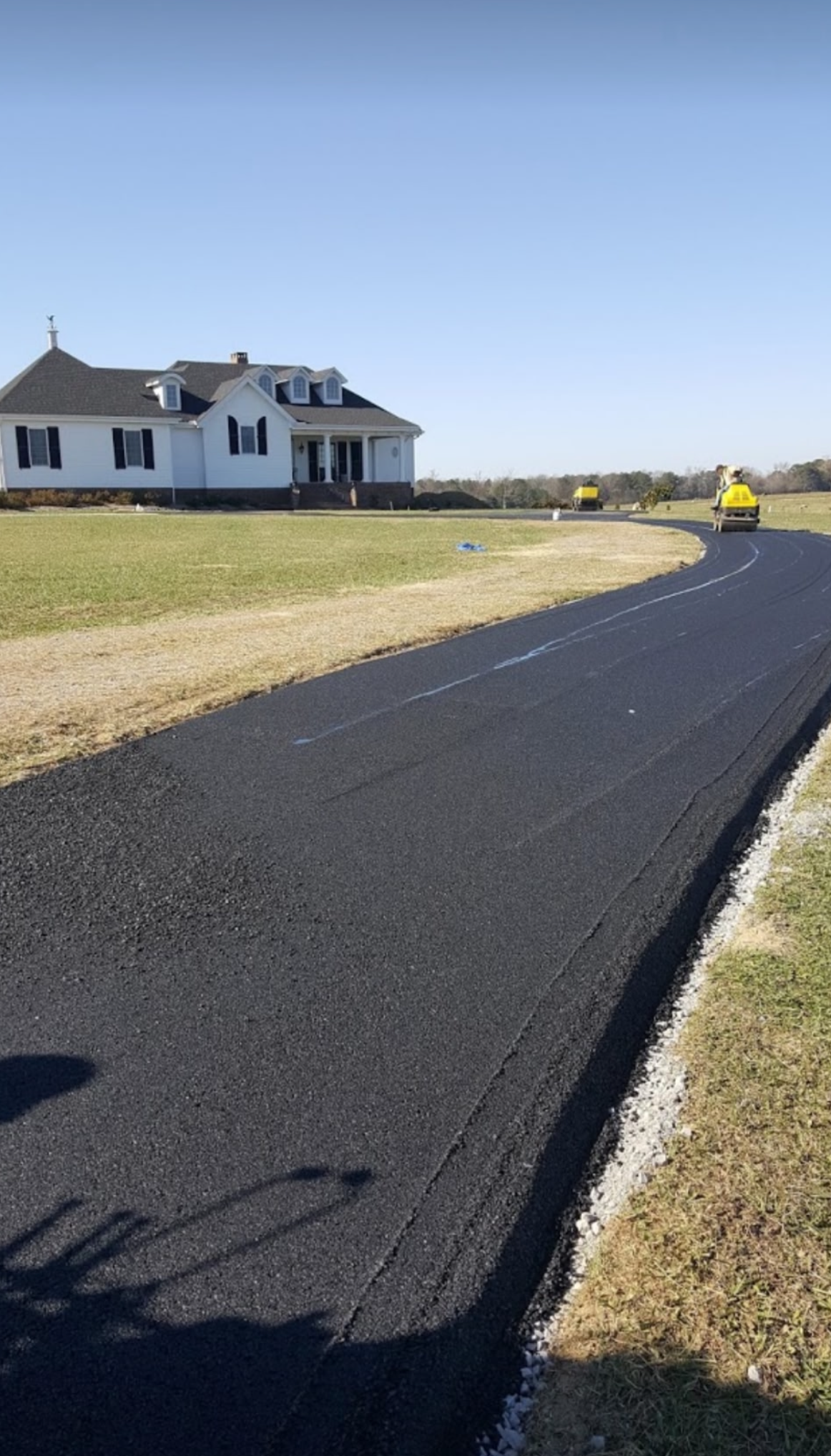 Newly paved asphalt driveway leading to a white house with black shutters, yellow construction equipment in the background.