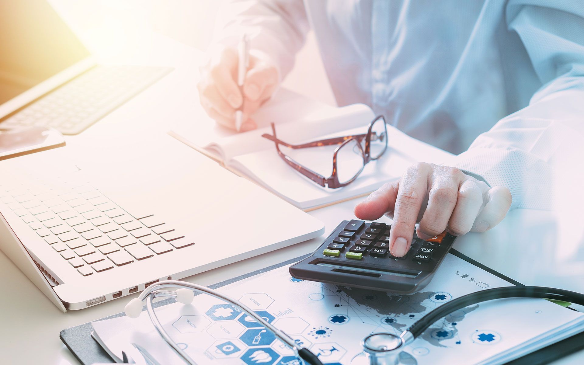 A doctor in a lab coat working at a desk, using a calculator and writing, with a stethoscope and medical documents.