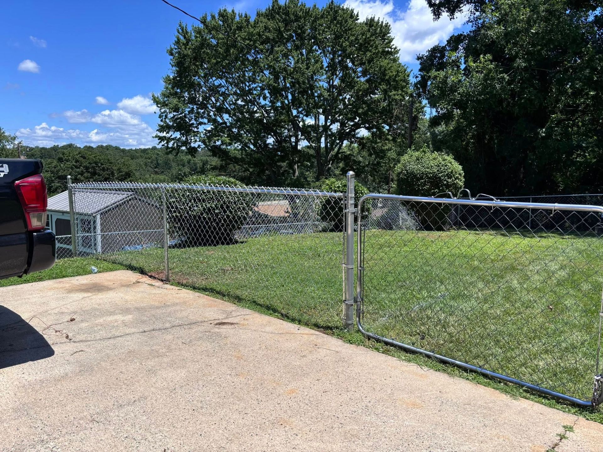 Chain-link gate beside a driveway opening into a grassy yard with trees and a blue sky