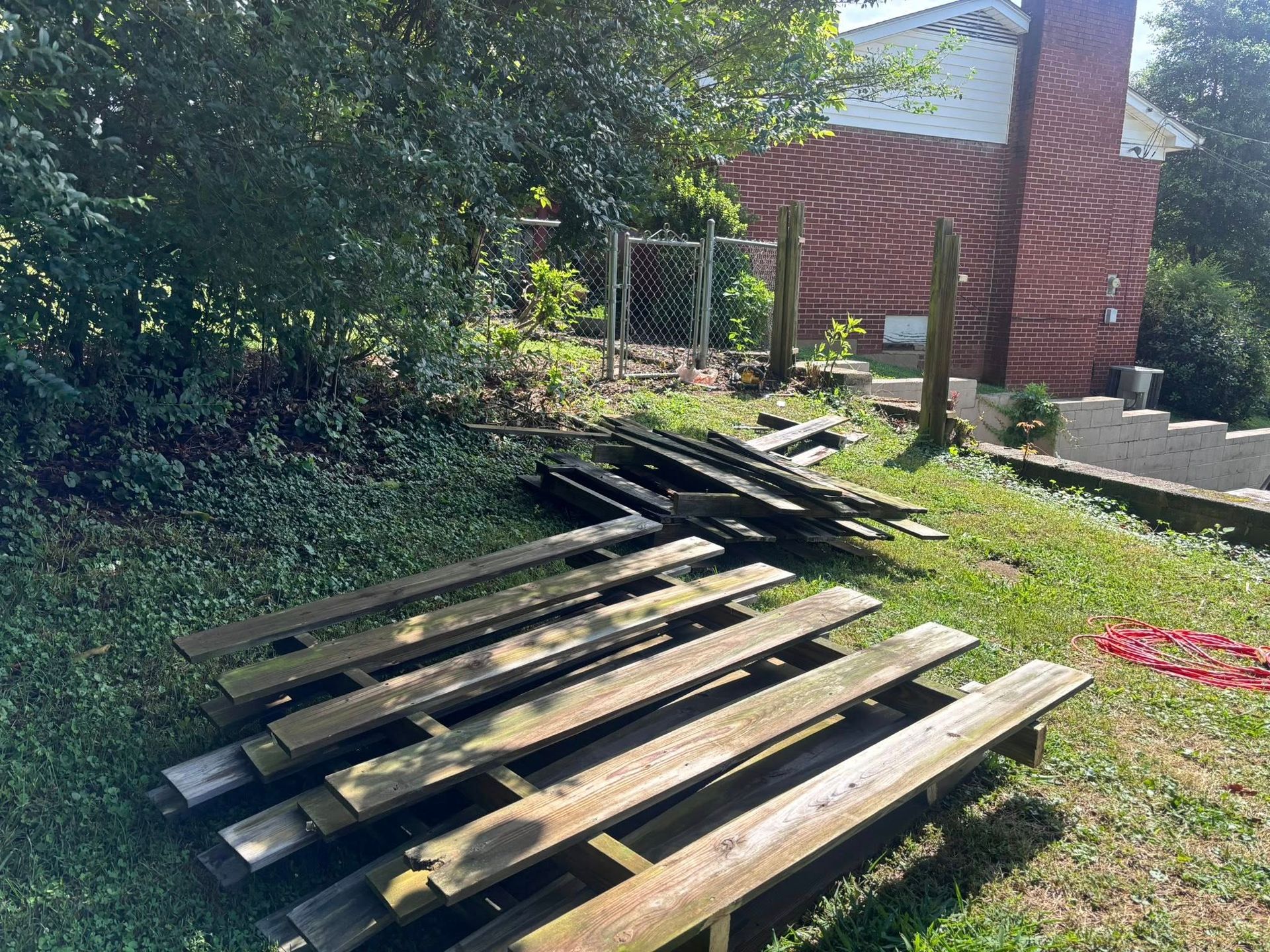 Stacked wooden pallets on a grassy slope beside a brick building and trees