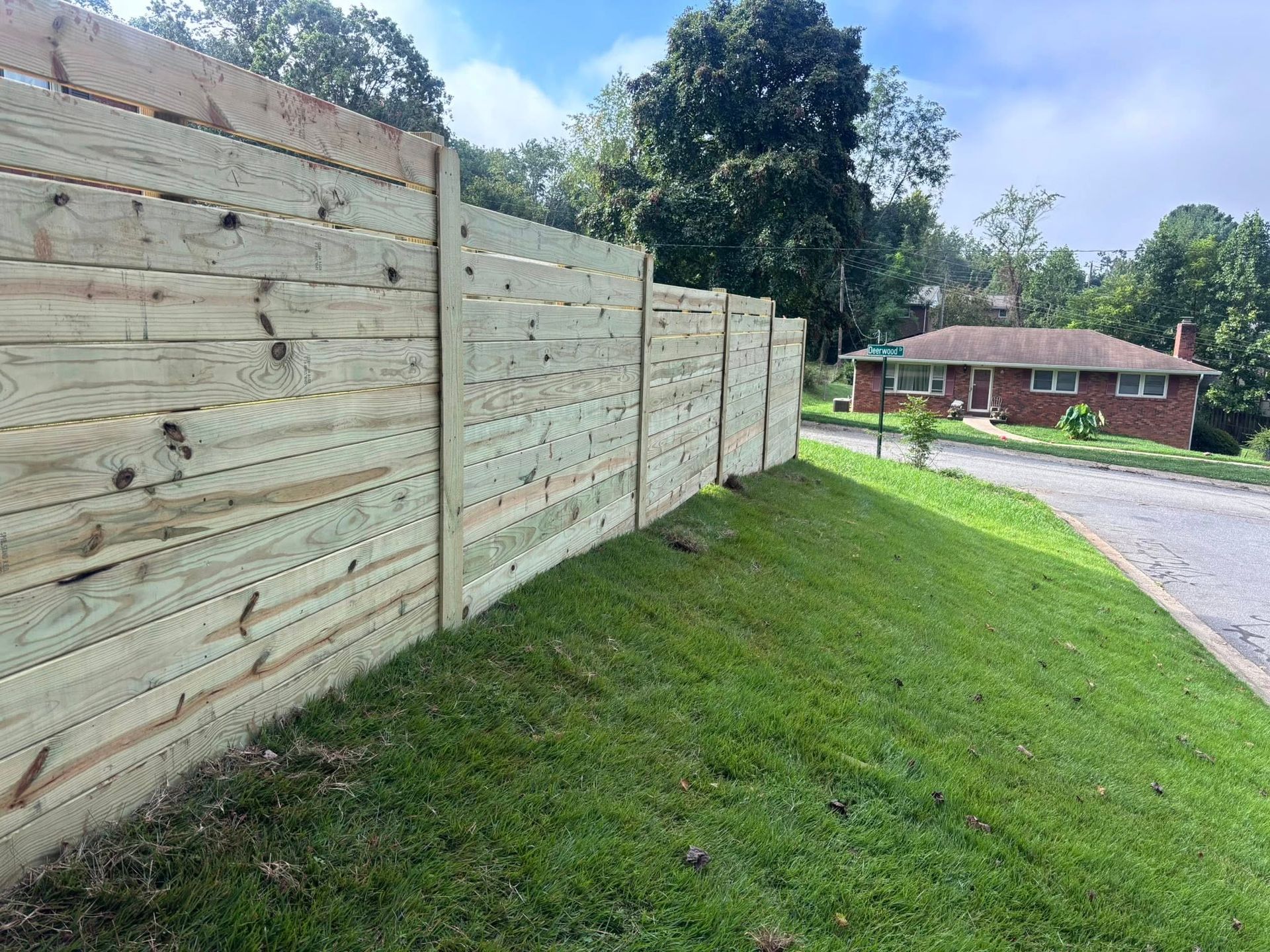 Wooden privacy fence beside a grassy slope and road, with a house in the background