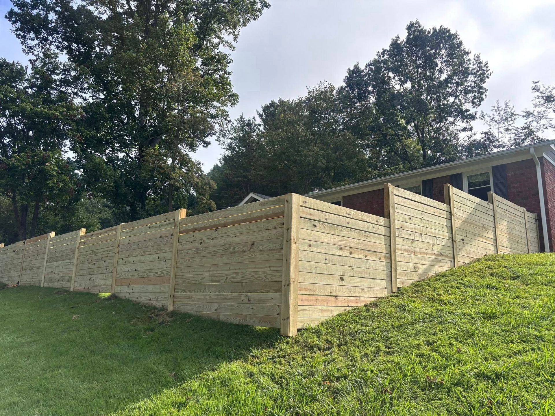 Wooden retaining wall on a grassy slope beneath tall trees and a cloudy sky
