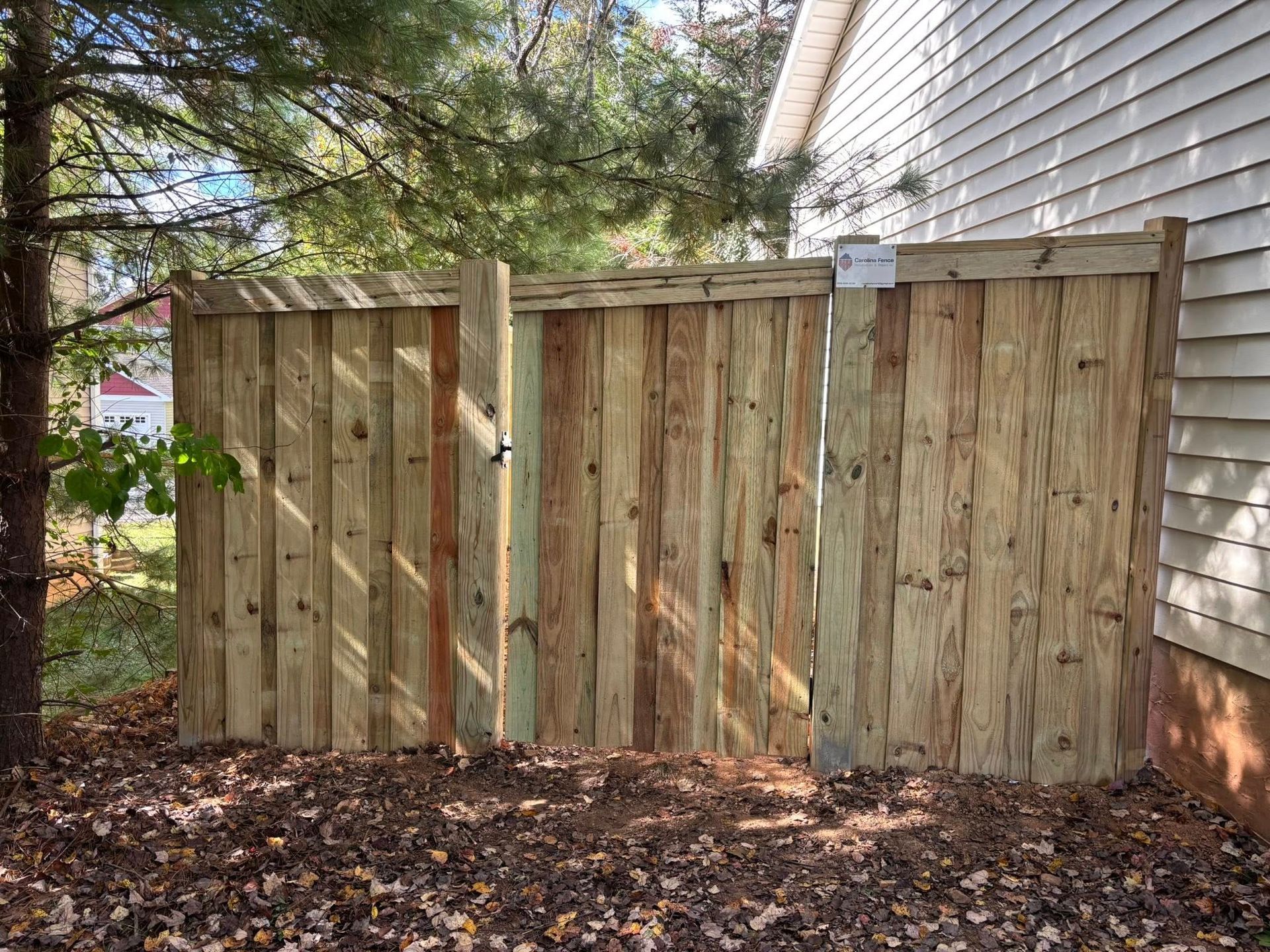 Wooden fence panels beside a house, with trees and fallen leaves in a yard.